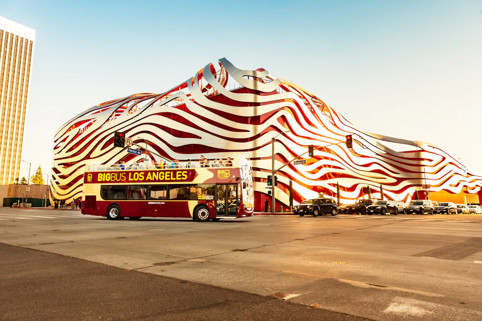 Double-decker bus labeled "Big Bus Los Angeles" on a city street with a wavy, red-and-white striped building in the background.