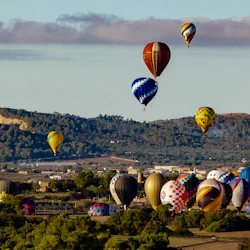 Hot air balloons in various colors and patterns floating over a hilly landscape with trees and buildings.