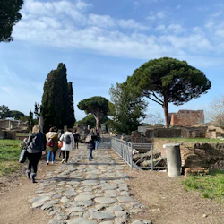 Människor som går på en stenväg genom antika ruiner med träd och blå himmel i bakgrunden.