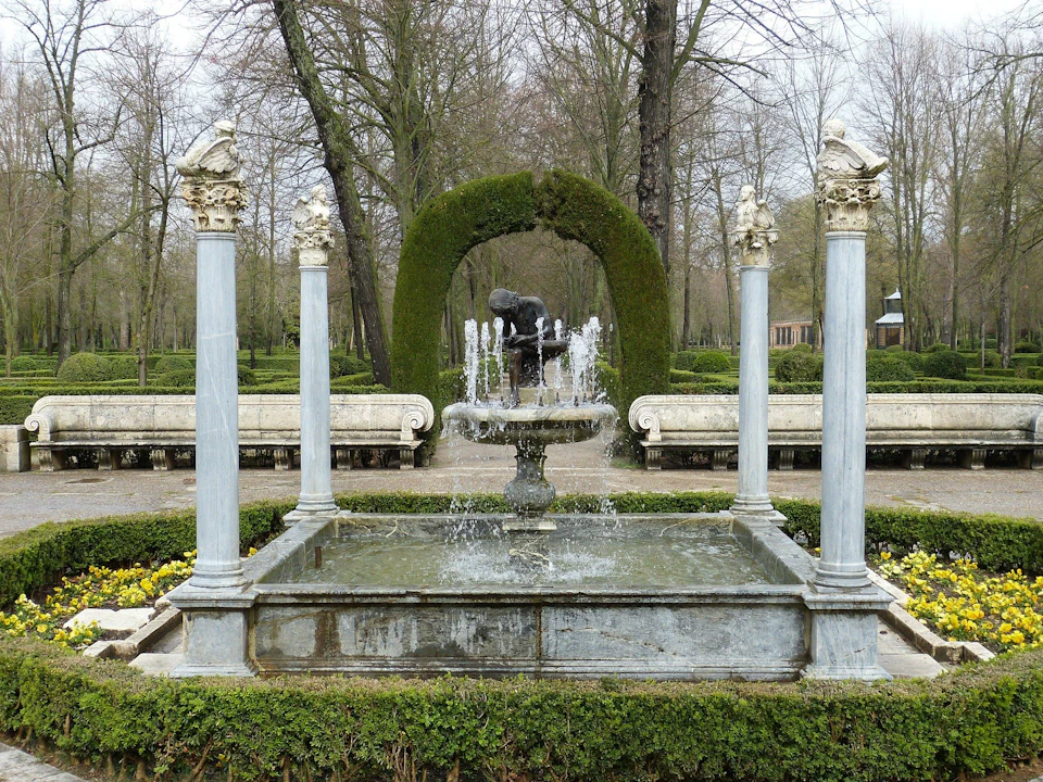 Stone fountain with four pillars and a bench in a park. An arched hedge and leafless trees are visible in the background.