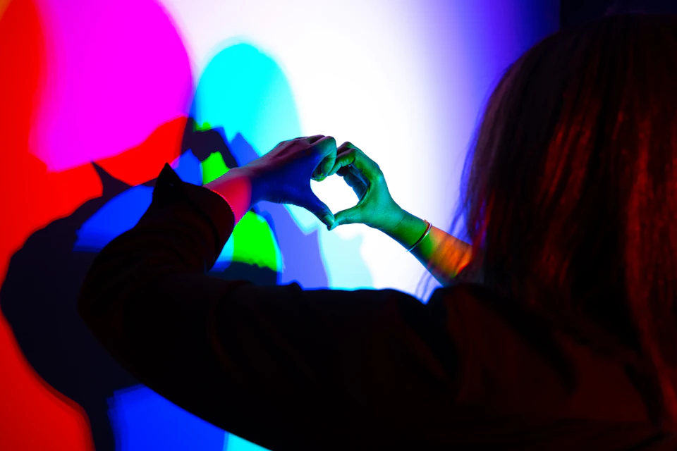 A person forming a heart shape with their hands in front of a colorful light projection creating overlapping shadows.