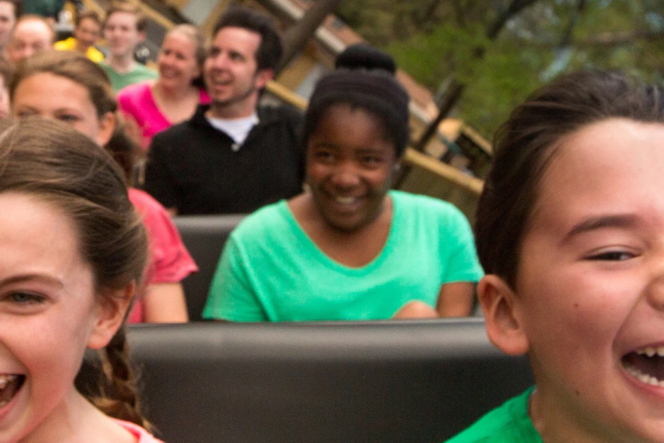 Two children in the front row of a wooden roller coaster smile with excitement. Other passengers and trees are visible in the background.