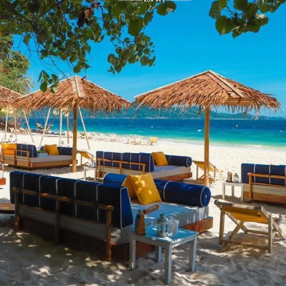 Beach setting with thatched umbrellas, blue cushioned lounge chairs, yellow pillows, and small tables. Blue sea and blue sky in the background.