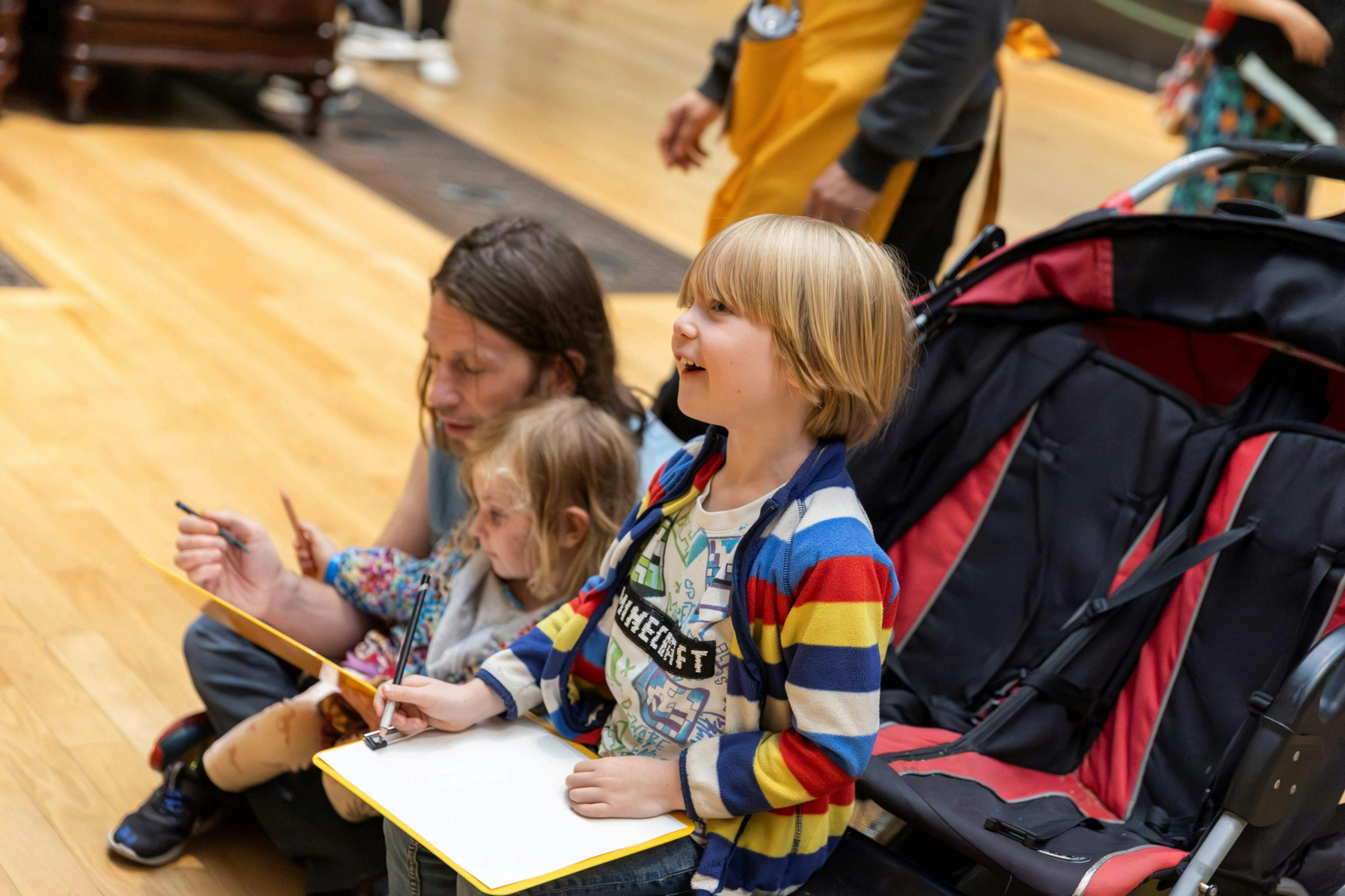Δύο παιδιά με σημειωματάρια και ένας ενήλικας που κάθεται σε ένα ξύλινο πάτωμα. Το ένα παιδί κρατάει ένα μολύβι, το άλλο χαμογελάει και ο ενήλικας κοιτάζει κάτω.