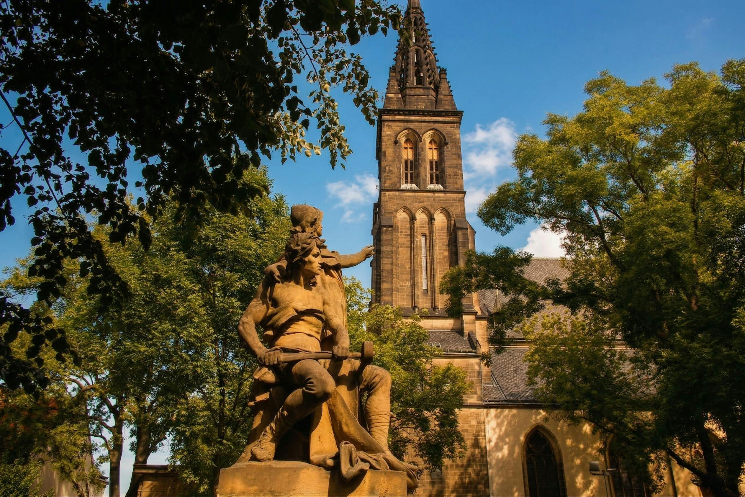 A statue in front of a tall, historic church tower surrounded by trees under a clear blue sky.