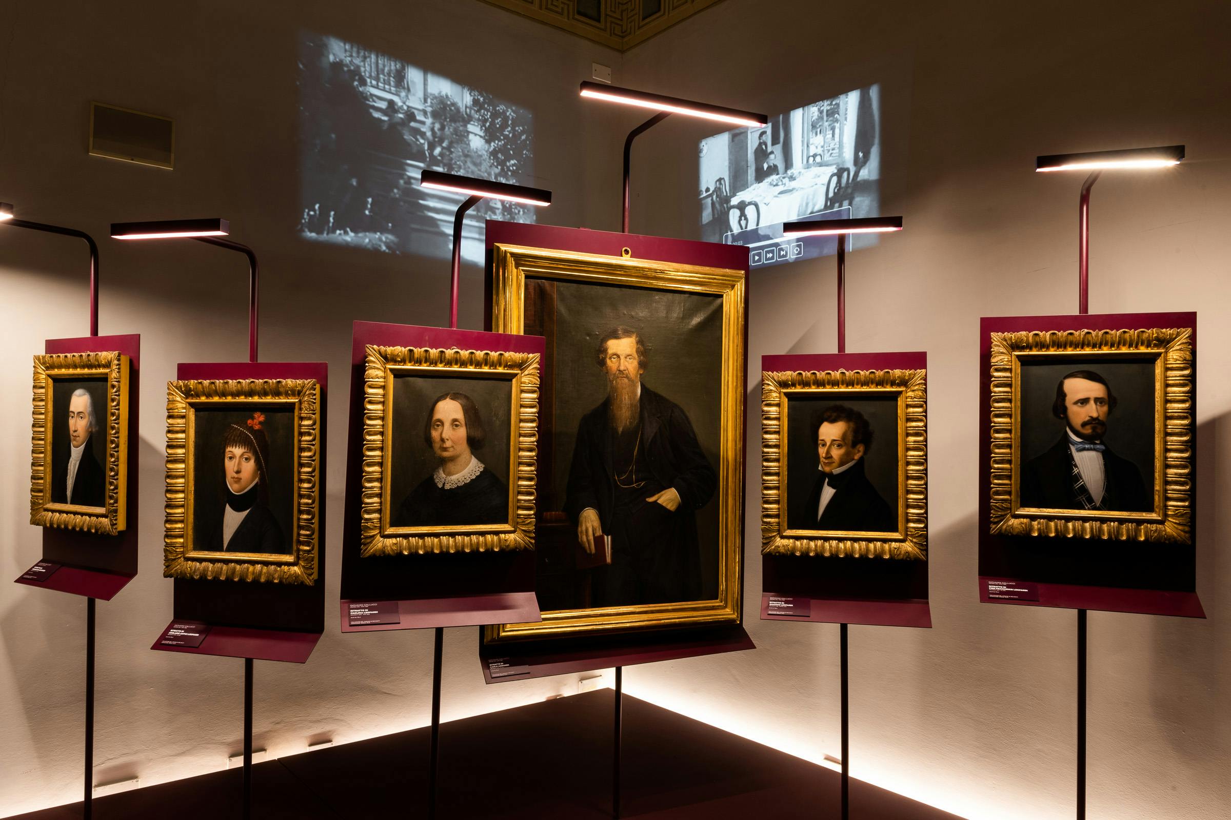 Ritratti incorniciati esposti in una galleria poco illuminata da luci moderne. Proiezioni di scene d'epoca sulle pareti.