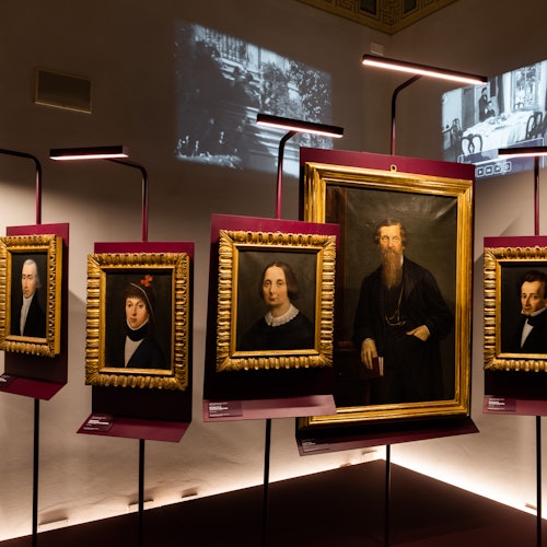 Framed portraits displayed in a dimly lit gallery, illuminated by modern lights. Projections of vintage scenes on the walls.