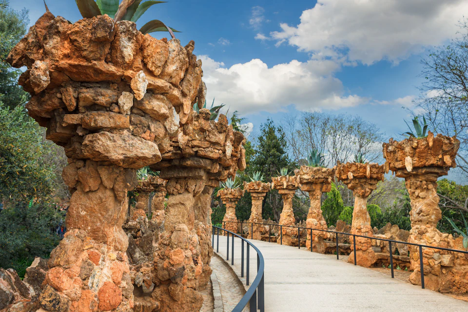Curved stone pathway lined with large, rocky columns topped with plants; blue sky with clouds in the background.