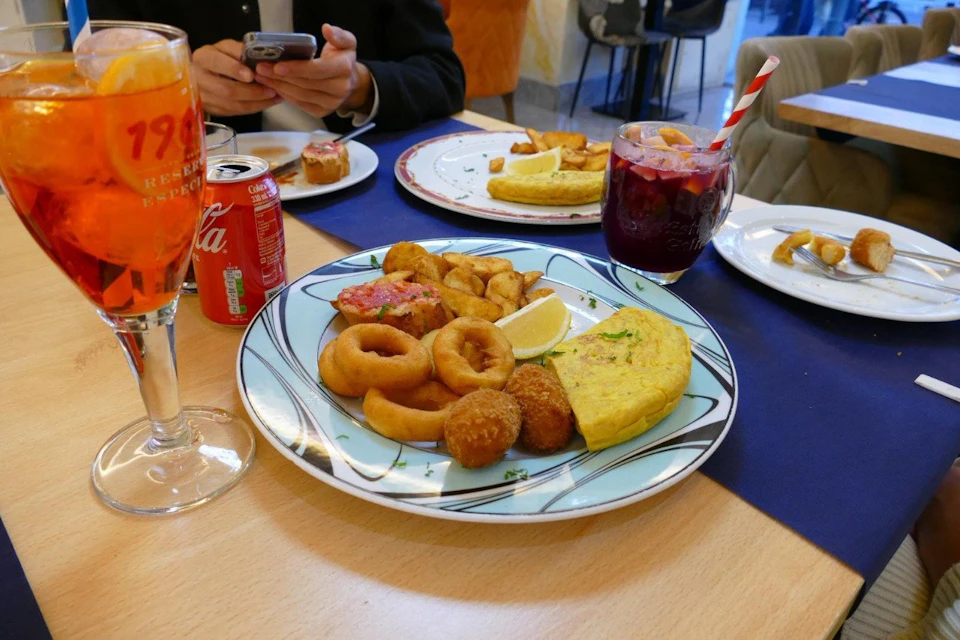 A table with plates of tapas, including tortilla, croquettes, and calamari, along with drinks like sangria and soda in a restaurant.