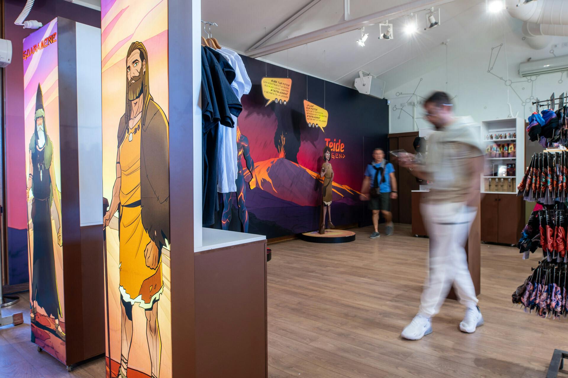 Et interiør i en butik med stativer af tøj og souvenirs, et vægmaleri på væggen og to slørede mennesker, der går forbi.