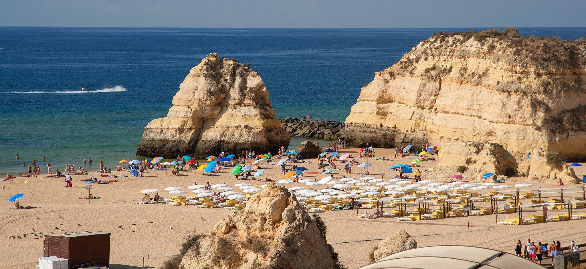 Crowded beach with colorful umbrellas and sunbathers, large rocky cliffs in the background, and a boat in the ocean.