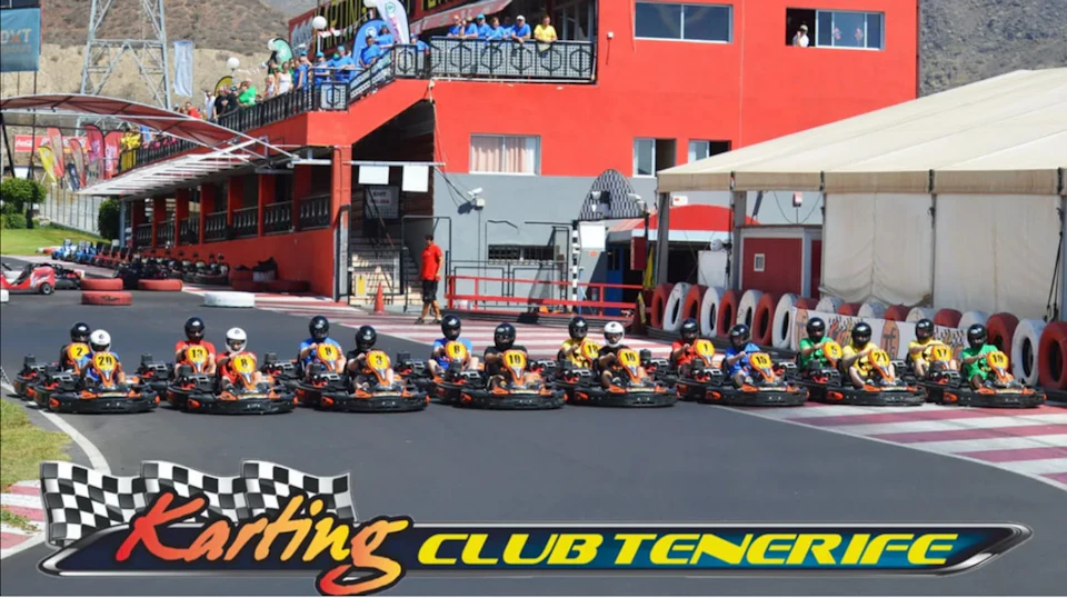A group of go-karts lined up on a track in front of a red building with spectators above and a sign reading 'Club Tenerife.'