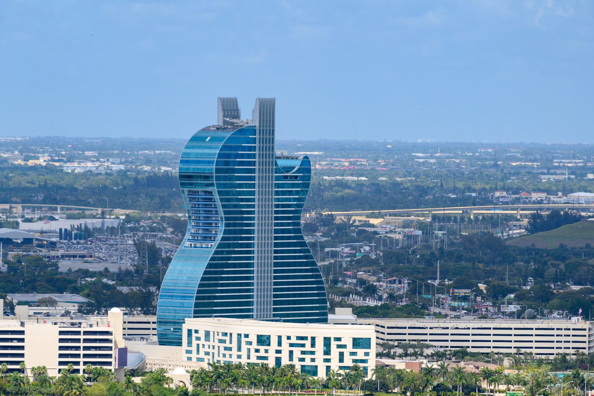 A large blue glass skyscraper shaped like a guitar, set in a cityscape with greenery and buildings in the background.