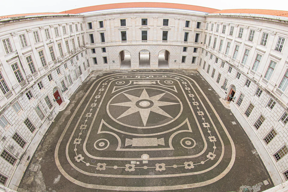 A large courtyard with geometric stone patterns is surrounded by a three-story building with arched entrances and windows.