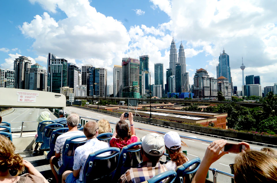 People on a double-decker bus tour overlooking a cityscape with modern skyscrapers and the Petronas Towers under a bright blue sky.