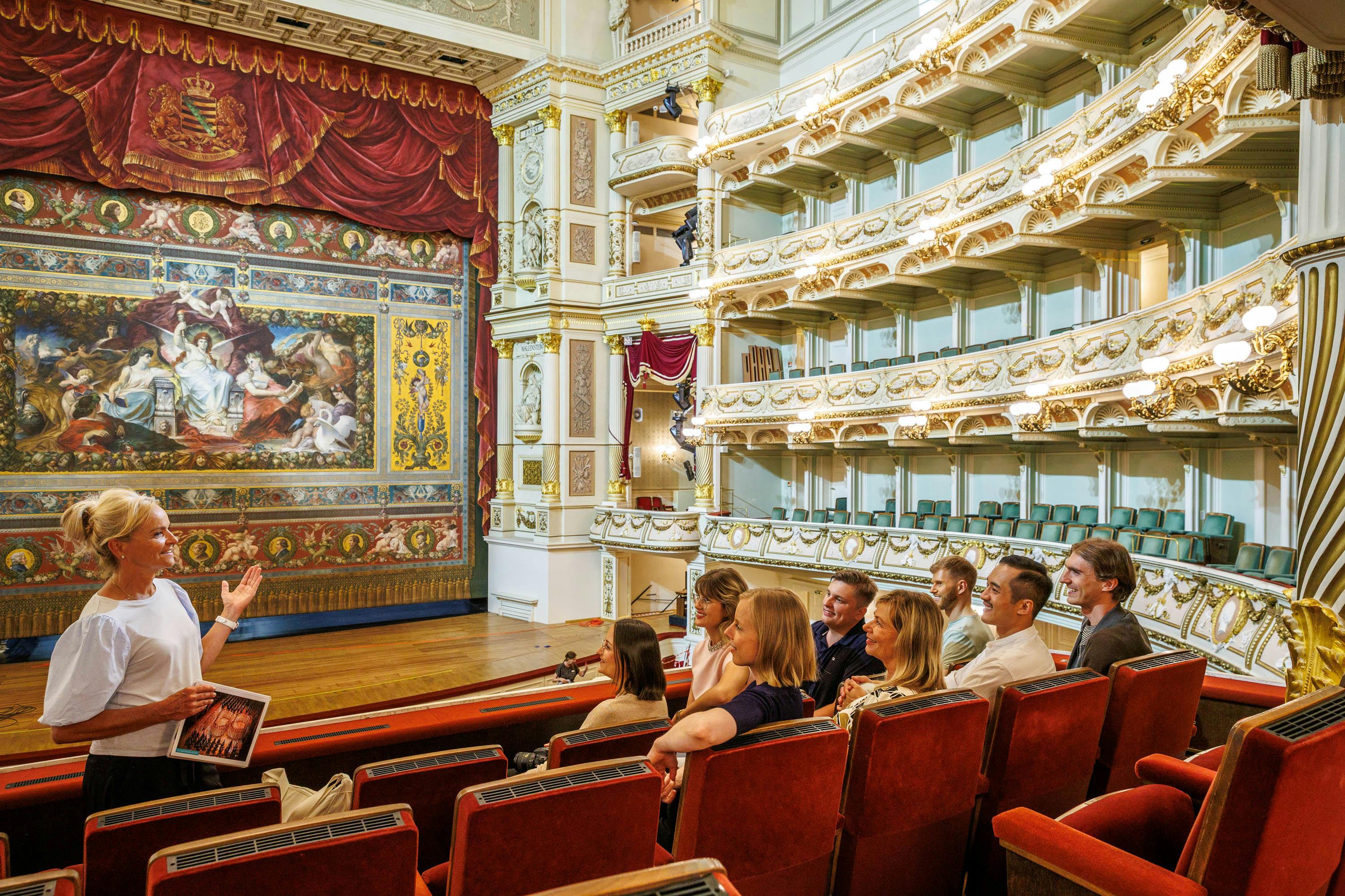 Un gruppo di persone sedute in un palco del teatro, che osservano l'interno riccamente decorato e a più livelli di un teatro dell'opera, con dettagli rossi e dorati.