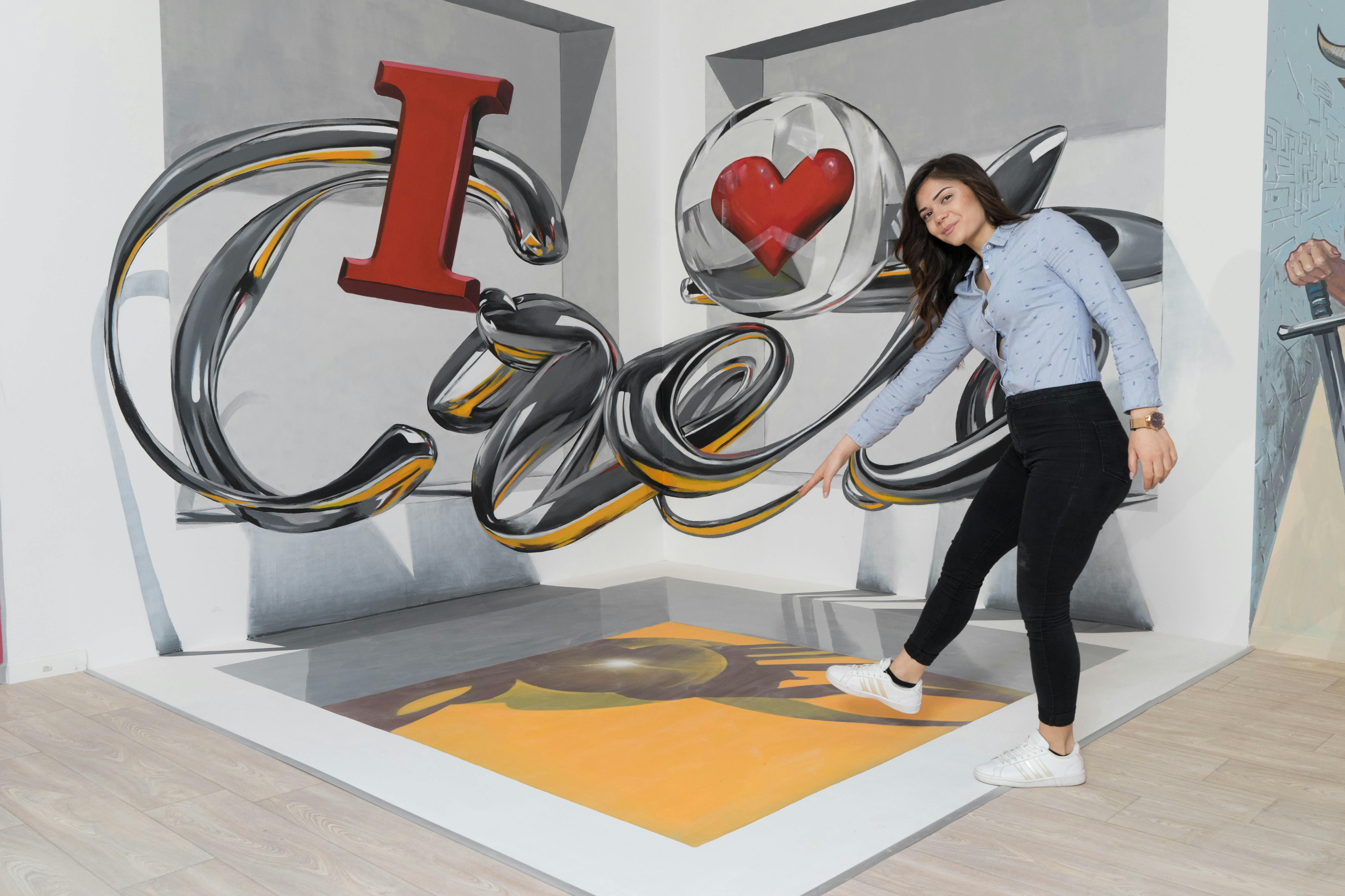 Žena stojící na 3D umělecké podlahové malbě s „I ❤️ Art“ na stěně, vytvářející iluzi hloubky.