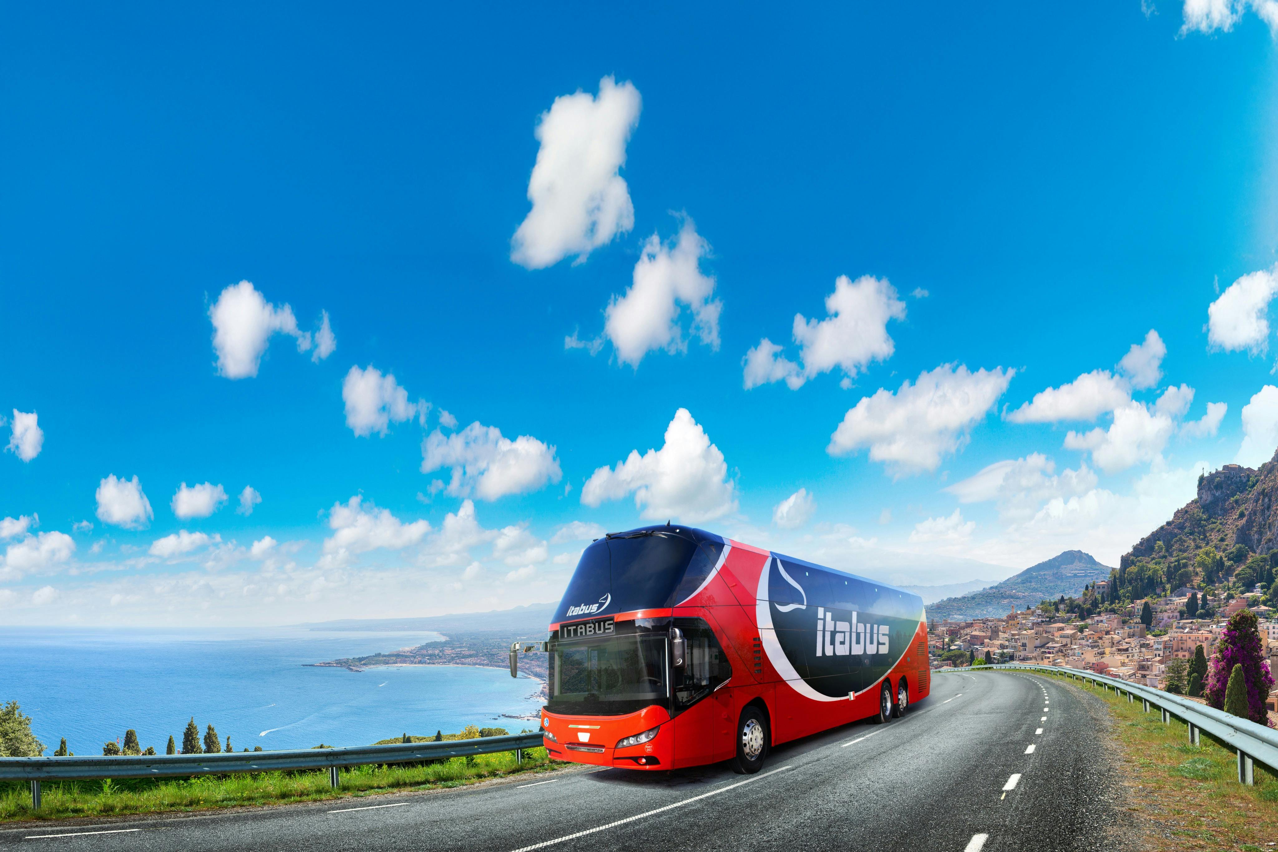 A red double-decker bus with "Itabus" branding travels on a scenic coastal road under a clear, blue sky with white clouds.
