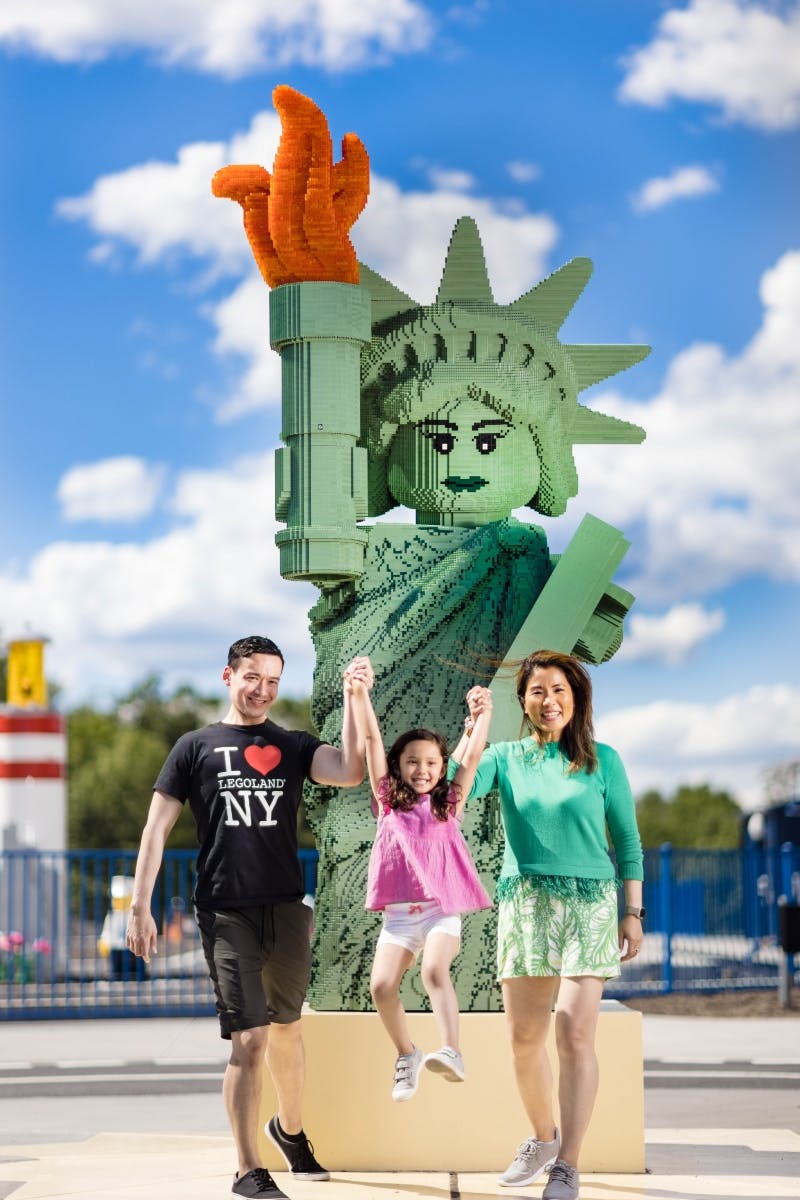 A smiling family holds hands with their daughter, lifting her in front of a LEGO Statue of Liberty.