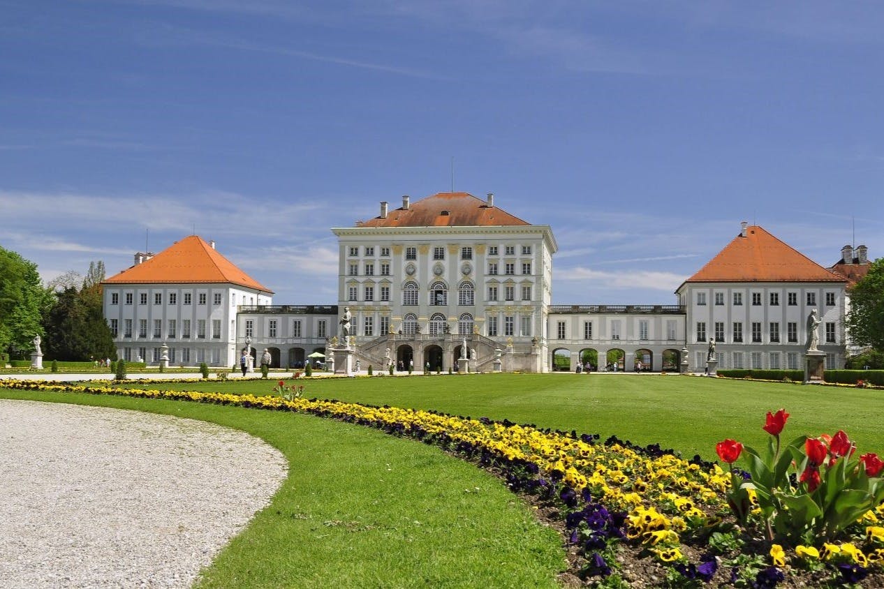 Gran palau blanc amb teulades vermelles, arquitectura ornamentada i jardins cuidados amb flors grogues i morades sota un cel blau clar.