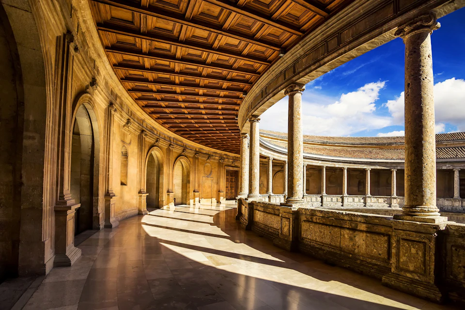 Sunlit circular stone corridor with carved arches and columns, wooden ceiling, and open sky view on the right. No people present.