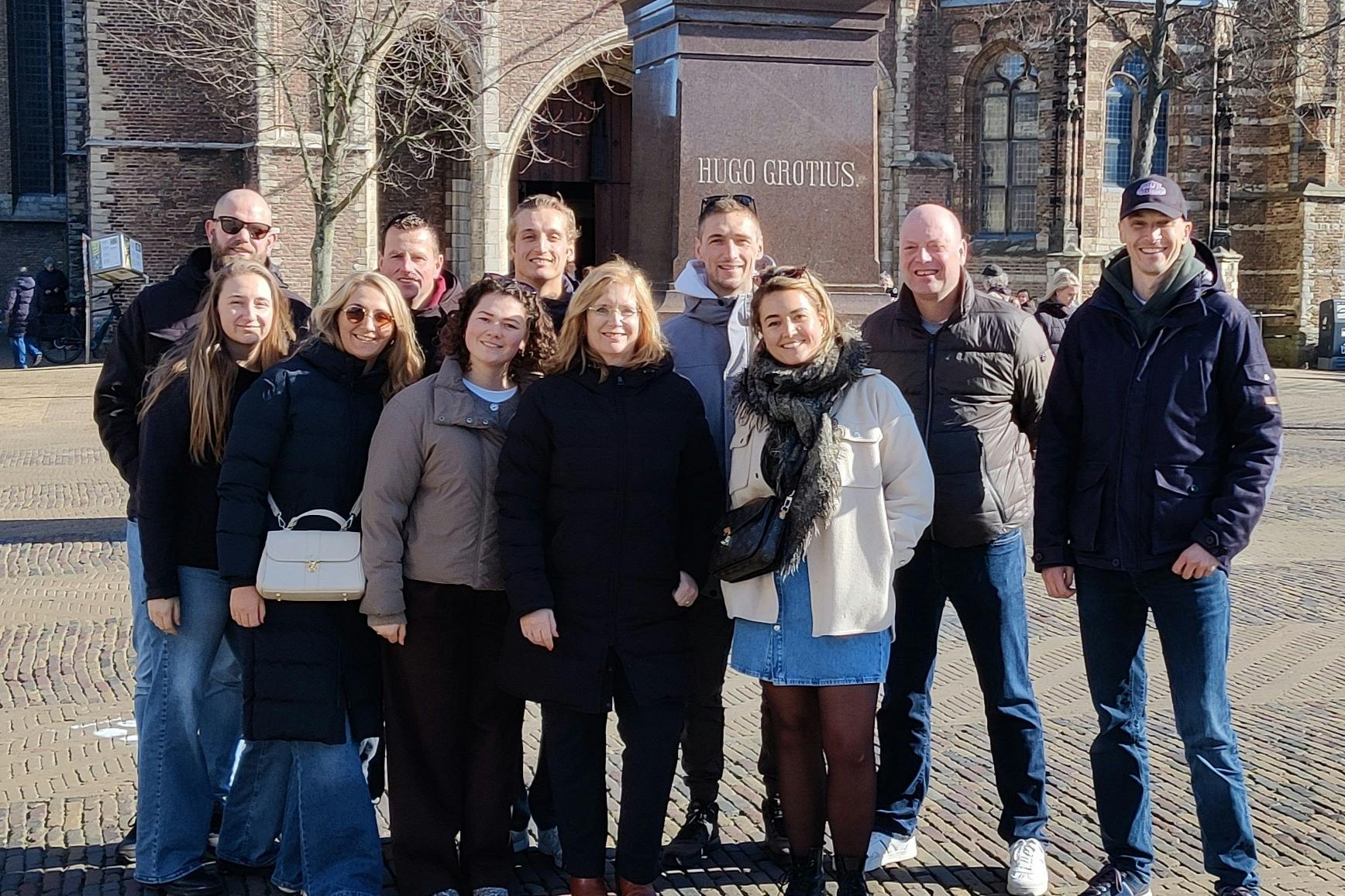 Skupina lidí pózující před sochou s nápisem „Hugo Grotius“ ve venkovním městském prostředí.