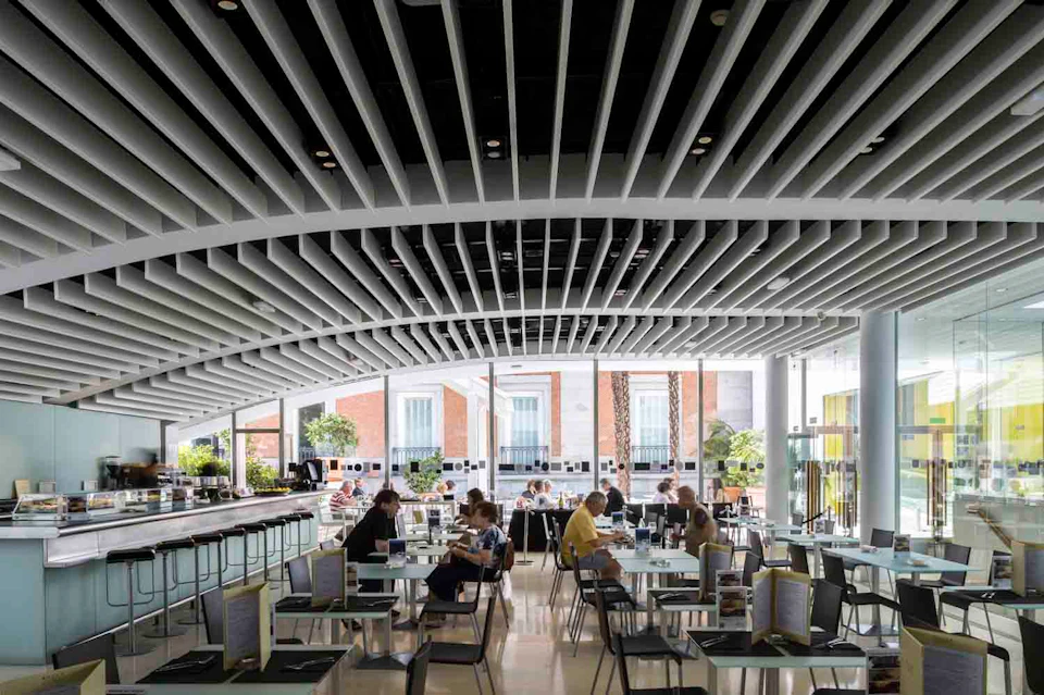 People sitting and dining at tables in a modern, well-lit cafeteria with large windows and a unique ceiling design.