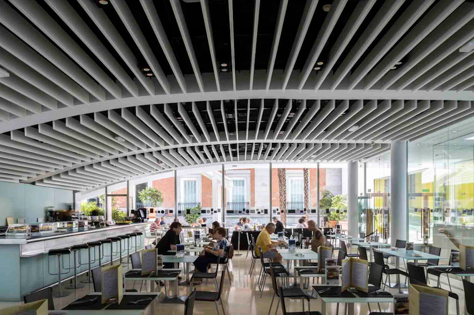 People sitting and dining at tables in a modern, well-lit cafeteria with large windows and a unique ceiling design.