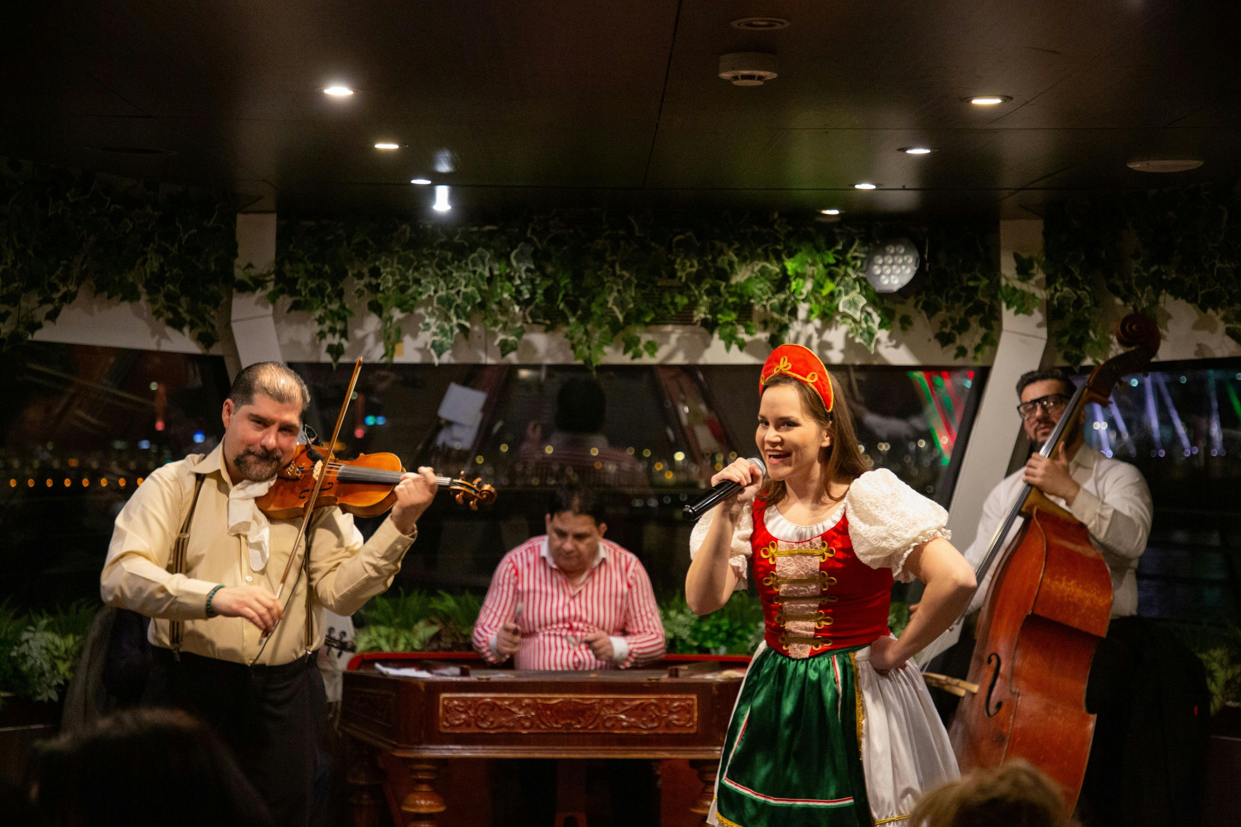 Женщина в традиционном наряде поет с живой группой, играющей на скрипке, контрабасе и забитом дульцимере в декорированной обстановке.