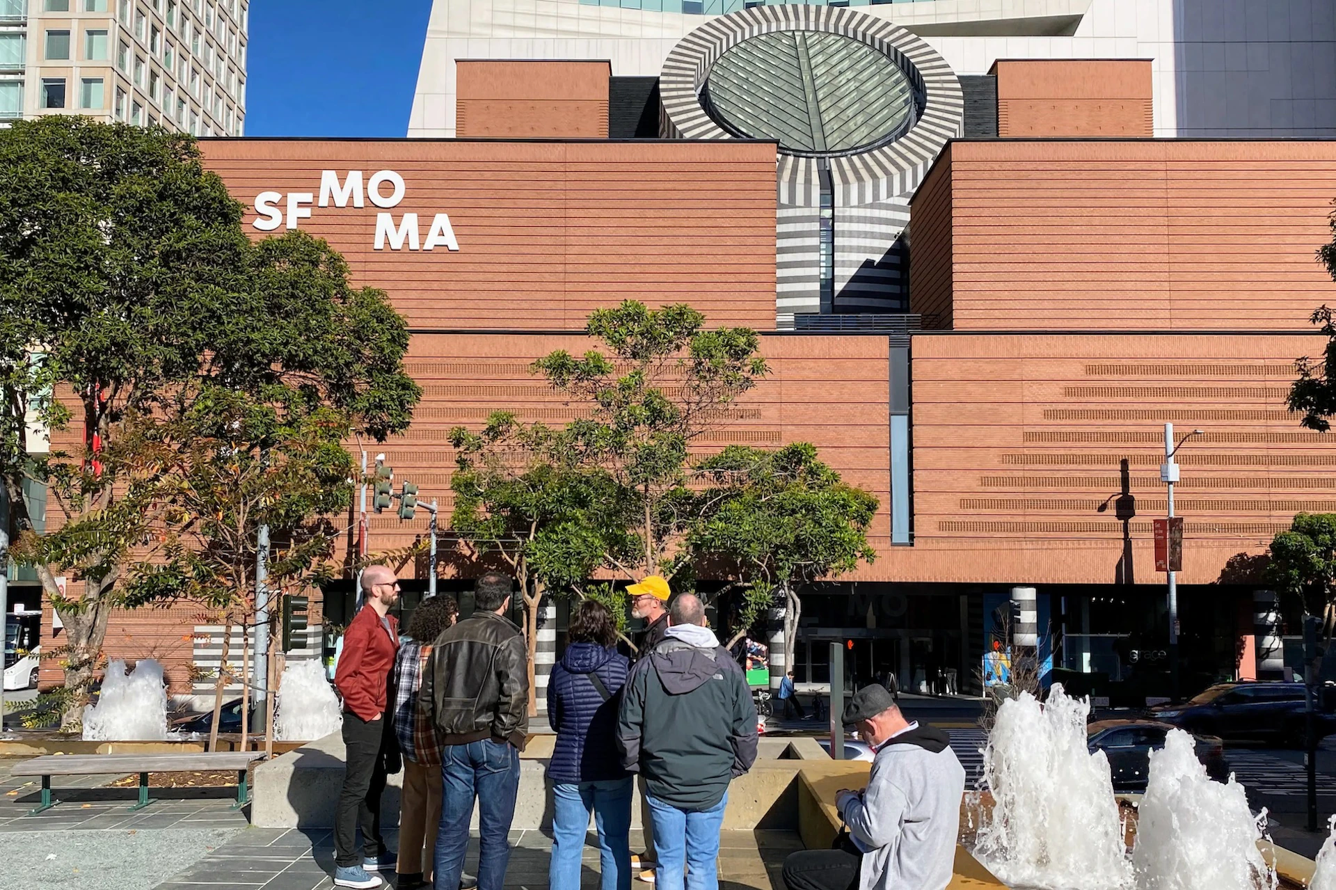 A group of people standing outside the SFMOMA building, characterized by its red brick facade, trees, and modern architectural elements.