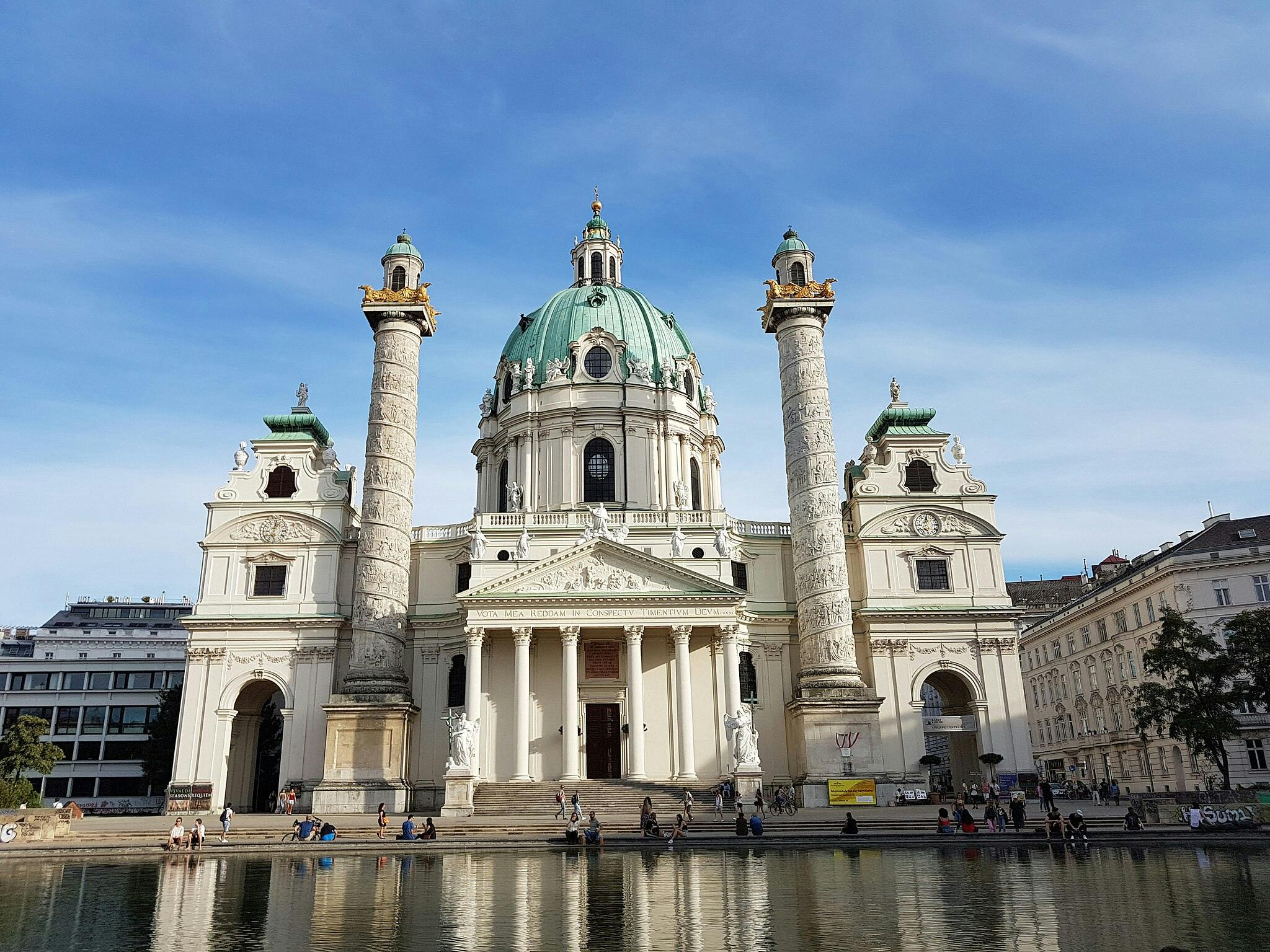 Barocke Kirche mit einer zentralen Kuppel, zwei verzierten Säulen und einem spiegelnden Becken davor. Die Menschen sitzen am Wasser.