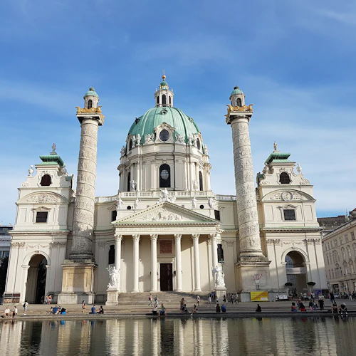Barocke Kirche mit einer zentralen Kuppel, zwei verzierten Säulen und einem spiegelnden Becken davor. Die Menschen sitzen am Wasser.