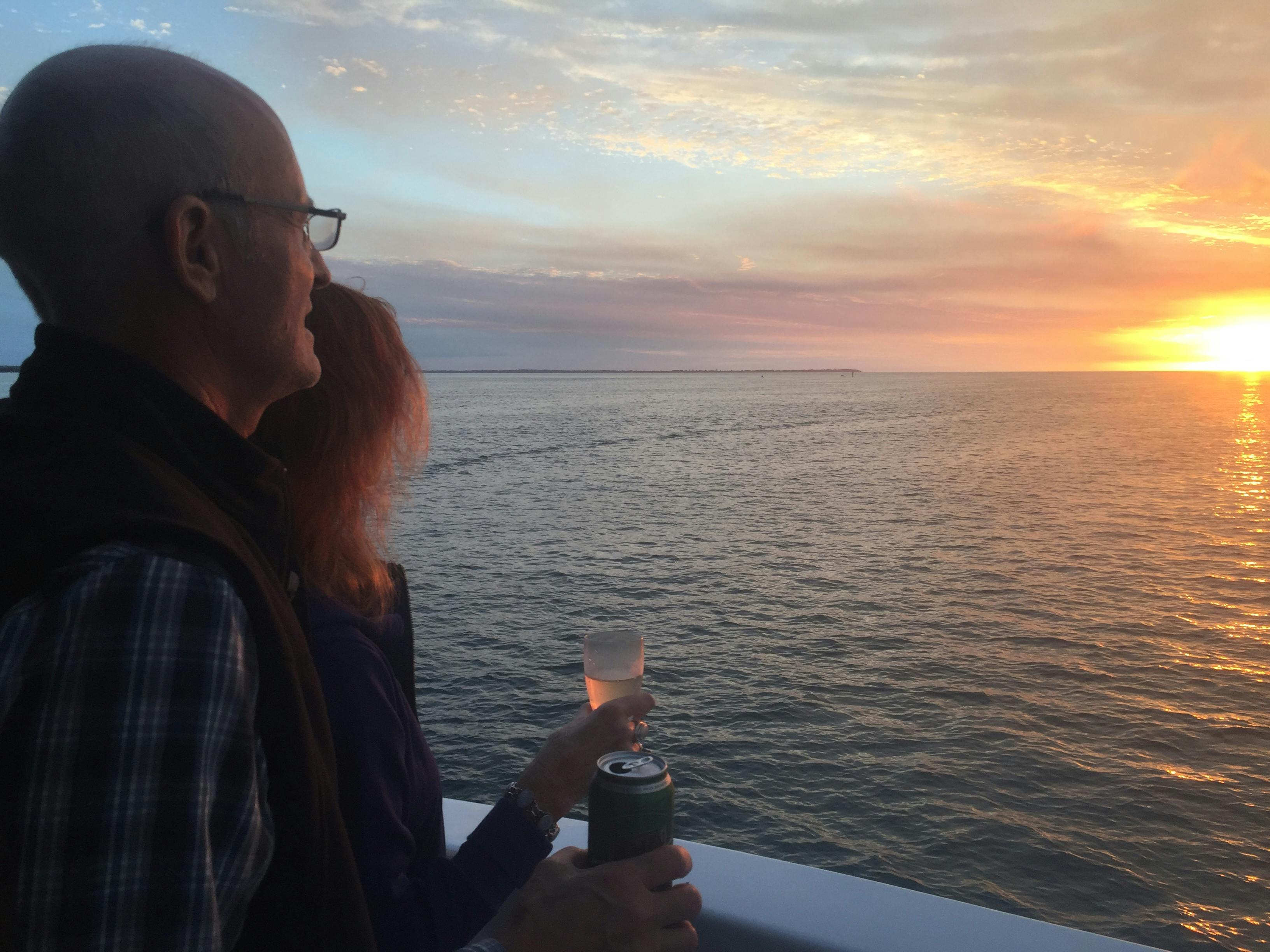 Two people on a boat deck, holding drinks, and watching a sunset over the ocean.