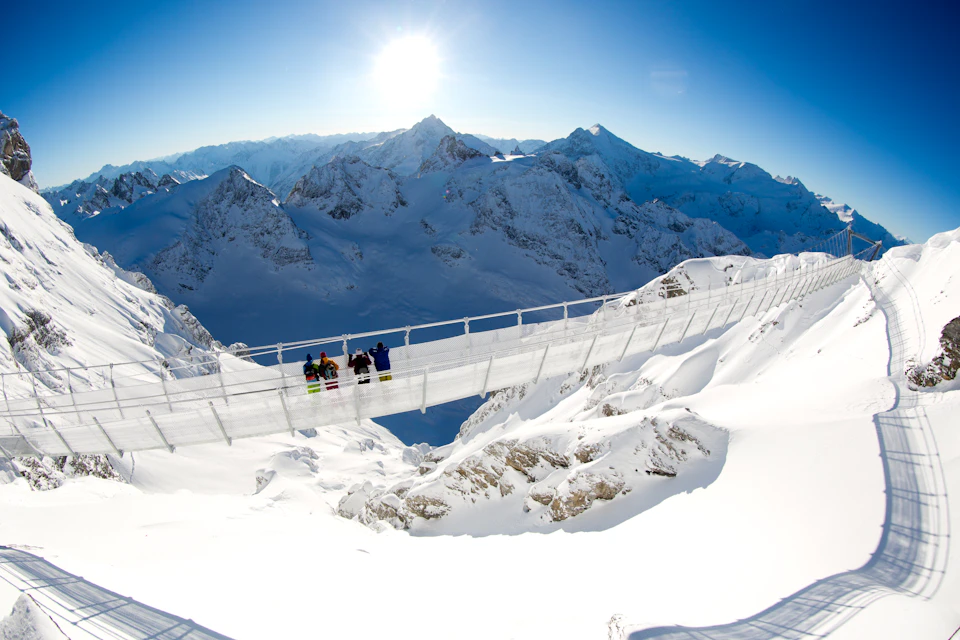 People stand on a suspension bridge over snowy mountains under a bright sun and clear blue sky.