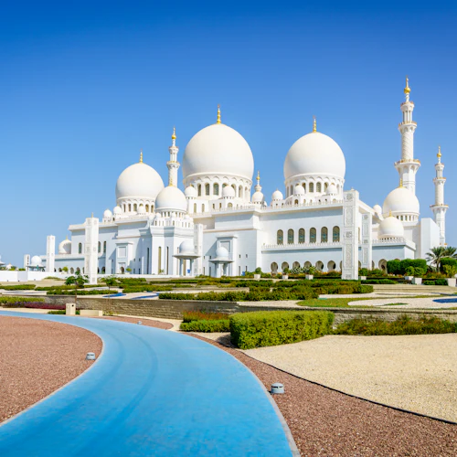Eine große weiße Moschee mit mehreren Kuppeln und Minaretten, umgeben von Gärten und einem blauen Weg unter einem klaren blauen Himmel.
