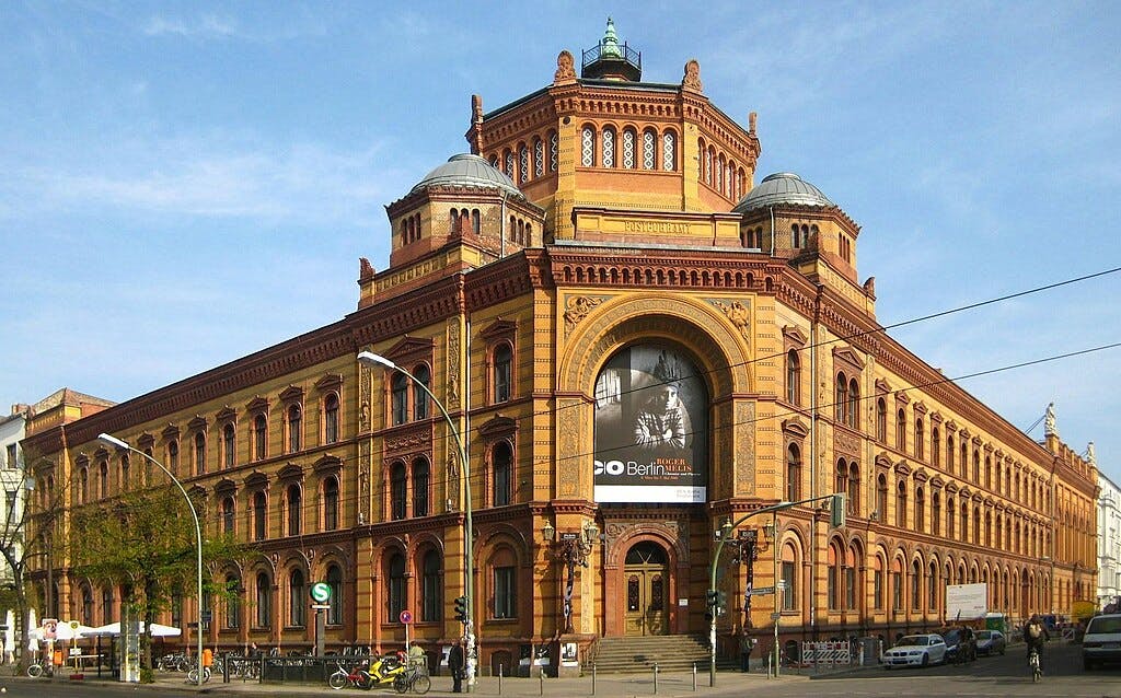 Large, historic brick building with arched windows and domed towers, located at a street corner with a visible poster on the facade.