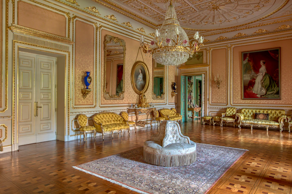 Opulent room with a large chandelier, ornate gold furniture, framed paintings, and a central cushioned seat on an intricate rug.