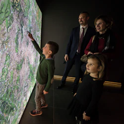 En pojke pekar på en gigantisk upplyst karta medan en leende man, kvinna och ung flicka observerar i ett mörkt rum.