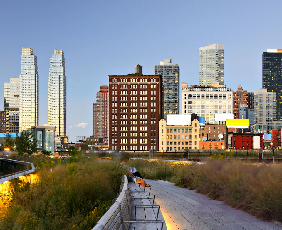 Pathway lined with greenery in the foreground, urban skyline with various buildings in the background at dusk.