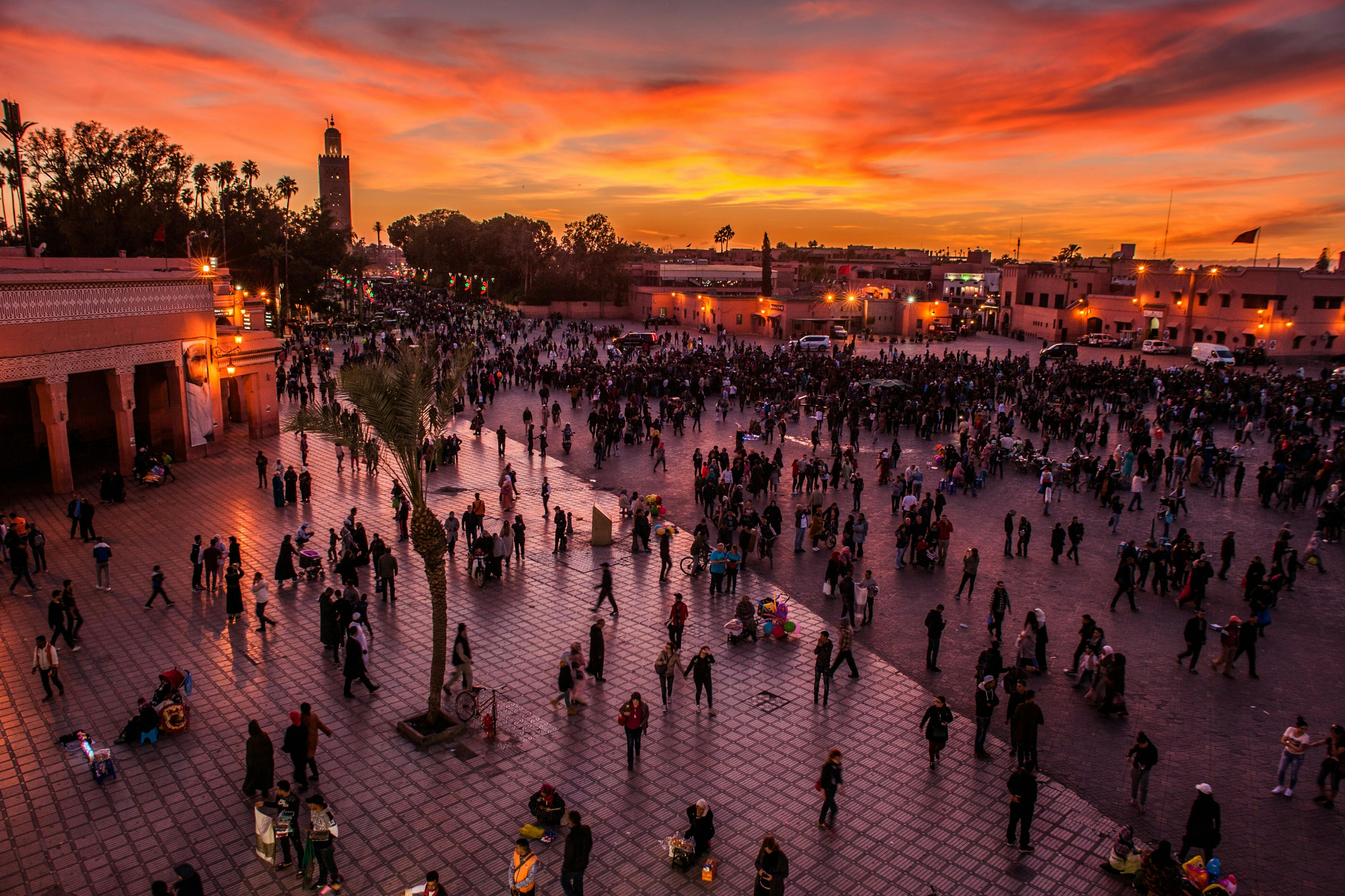 日落时分，人群聚集在一个大广场上，温暖的路灯照亮了广场，后面有尖塔和建筑物。