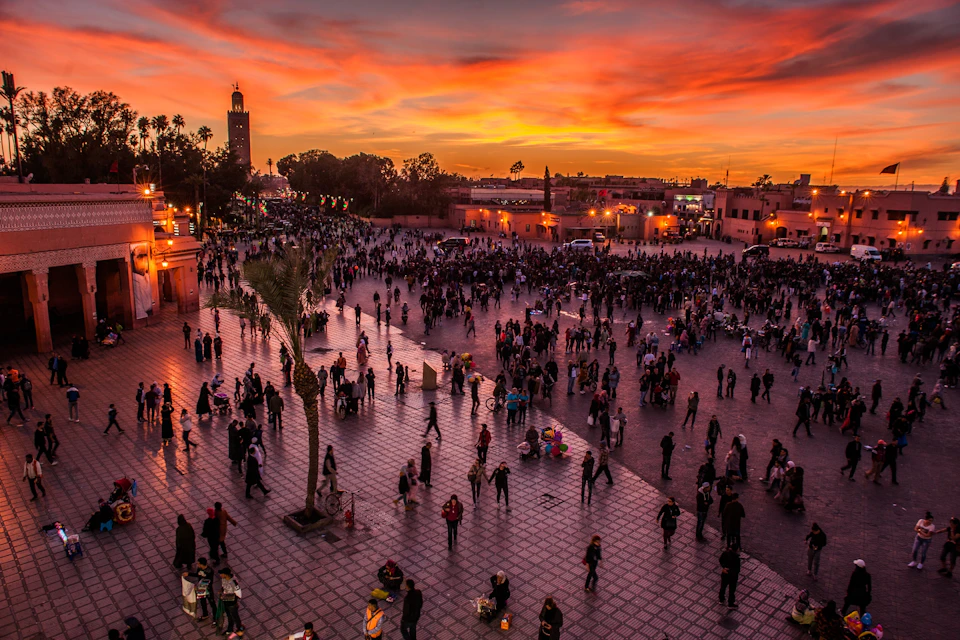 Crowds of people gather in a large plaza at sunset, illuminated by warm streetlights, with a minaret and buildings in the background.