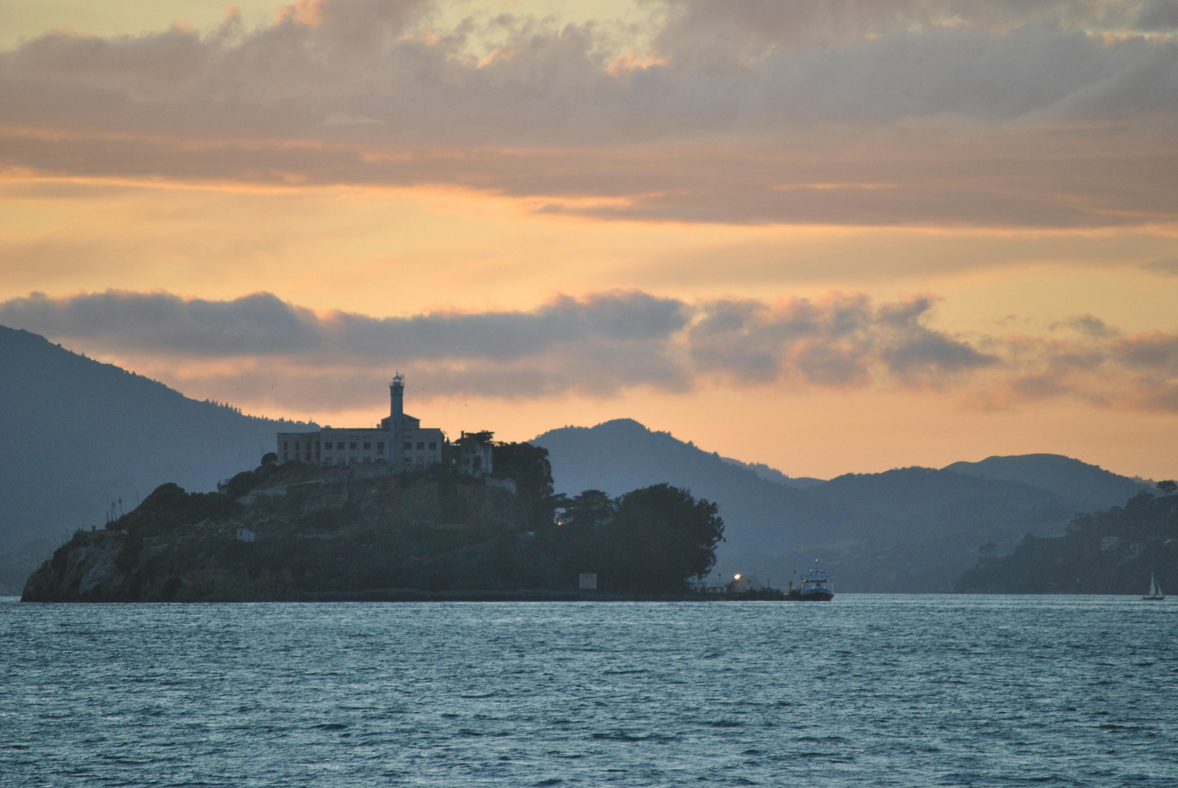 Uma ilha com um grande edifício e um farol, em silhueta contra um céu ao pôr do sol, rodeada por águas calmas e colinas distantes.