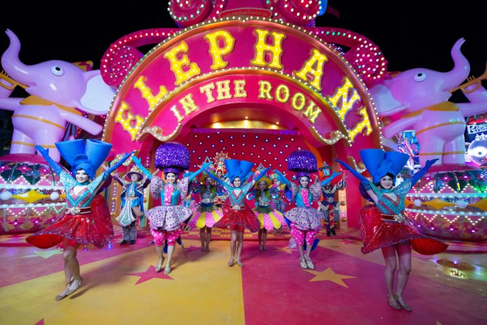 Performers in colorful costumes dance in front of a large sign reading "Elephant in the Room" with a pink elephant statue behind.