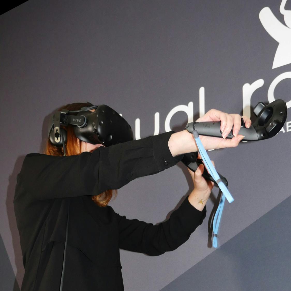 Person wearing a VR headset and holding a VR controller, standing in front of a wall with partial text and a logo.
