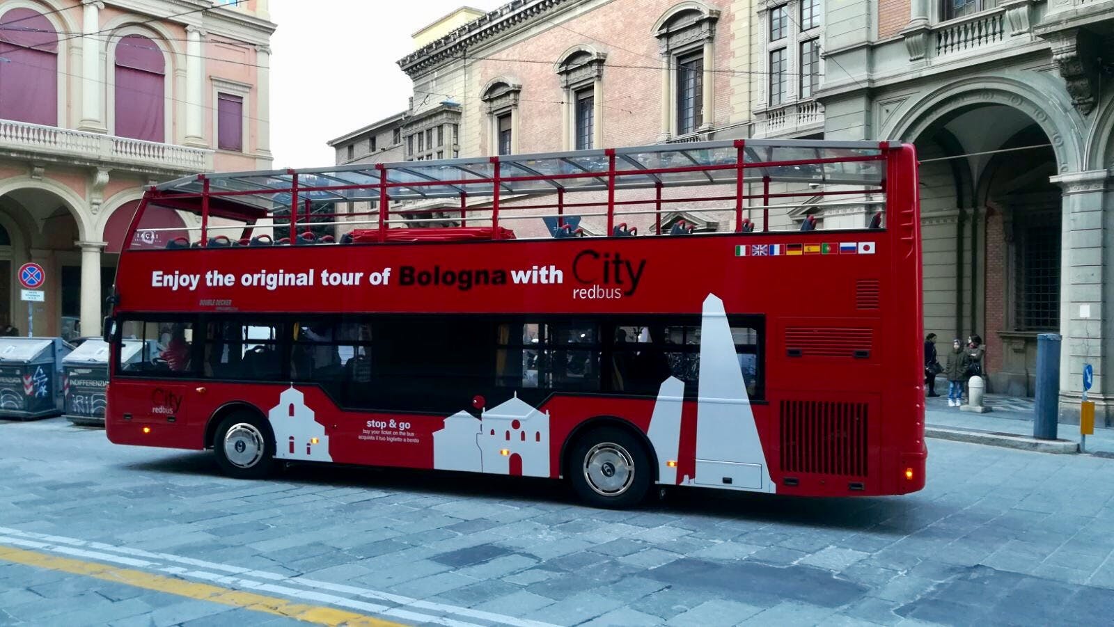 Rode dubbeldekker tourbus in Bologna, Italië, geparkeerd voor historische gebouwen. Tekst op de bus luidt &#34;Geniet van de originele stadstour door Bologna met City redbus.&#34;