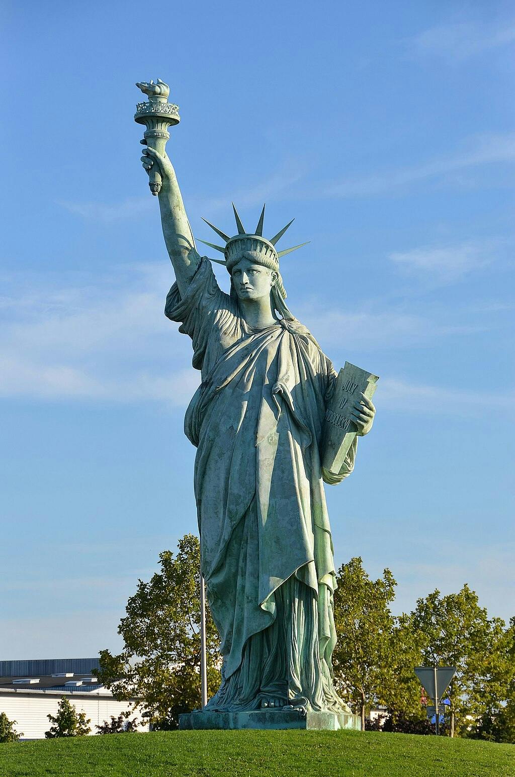 A statue resembling the Statue of Liberty, holding a torch and a tablet, stands on a grassy area under a clear blue sky.