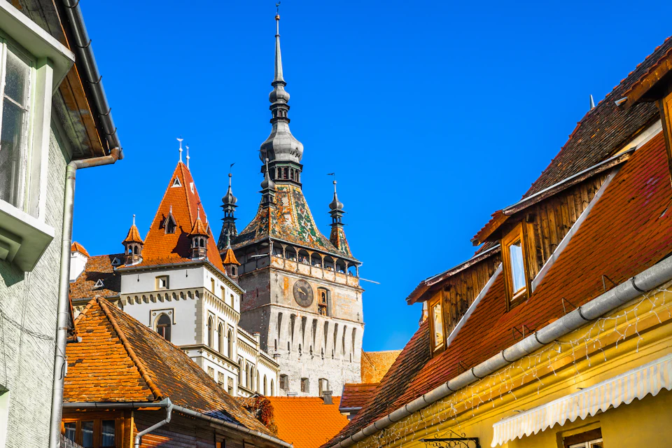 Historic clock tower with spires and colorful rooftops under a bright blue sky, viewed from a cobblestone street.