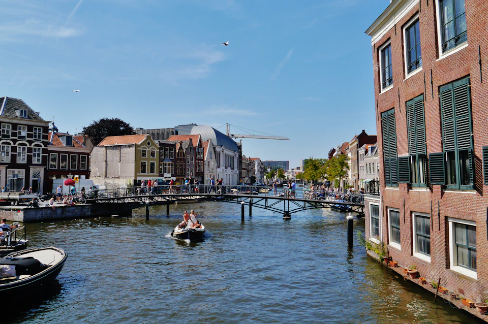 A canal with a boat, a bridge with pedestrians, historic buildings, and a crane in the background under a clear sky.