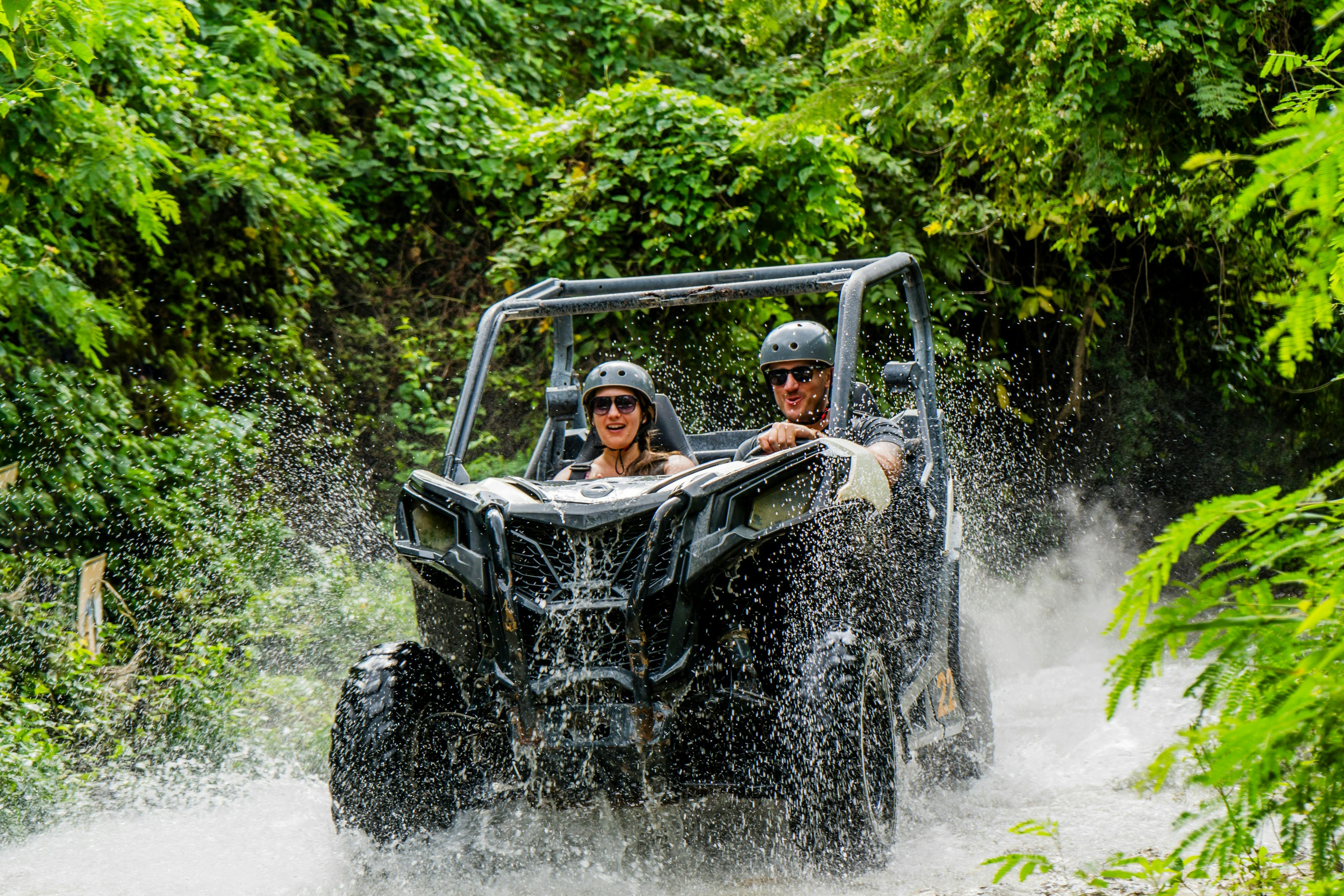 Two people wearing helmets drive an off-road vehicle through a shallow stream surrounded by lush greenery.