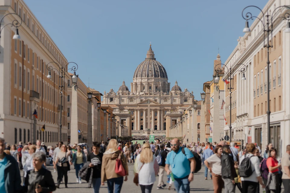 A crowded street in front of St. Peter's Basilica in Vatican City, with people walking and buildings lining the sides.